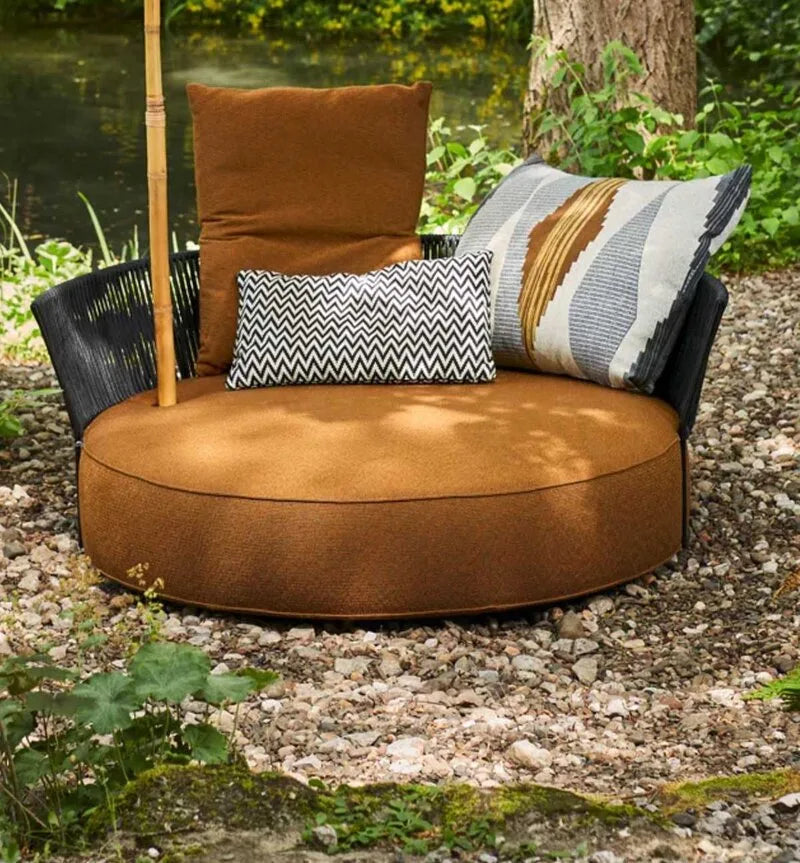 Bullfrog runder Outdoor-Loungesessel in cognacfarbenem Stoff mit gemusterten Kissen, schwarzem Geflecht und Bambusstütze in naturnaher Gartenumgebung.