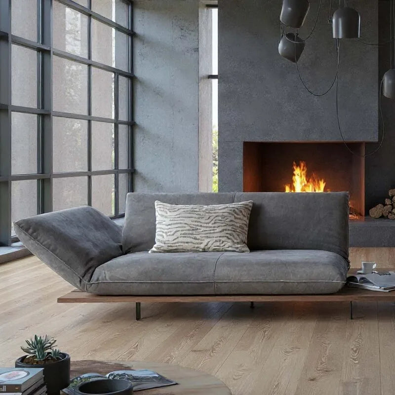 Bullfrog Sofa in Grau mit Lehnkissen vor Kamin, Betonwand und Sprossenfenster in modernem Loft-Wohnzimmer.