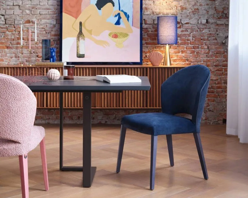 Mobitec Stühle in Rosa-Bouclé und dunklem Samtblau an einem schwarzen Tisch vor Backsteinwand mit Holzsideboard.