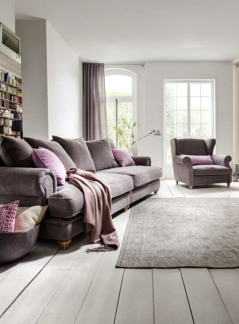 Helles Wohnzimmer mit graulila Polstersofa, passendem Ohrensessel, rosa Kissen, Plaid und Vintage-Teppich auf weißen Holzdielen.