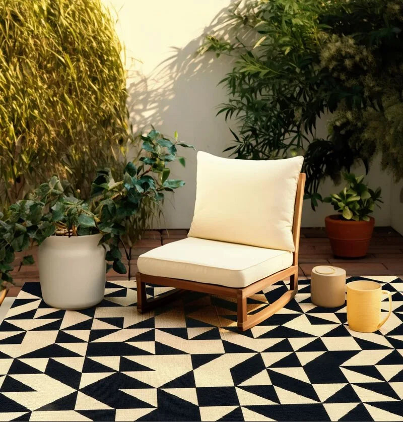 Schwarz-weißer Outdoor-Teppich mit geometrischem Dreiecksmuster auf einer sonnigen Terrasse, dazu Holz-Loungesessel mit cremefarbenen Polstern und Pflanzen.