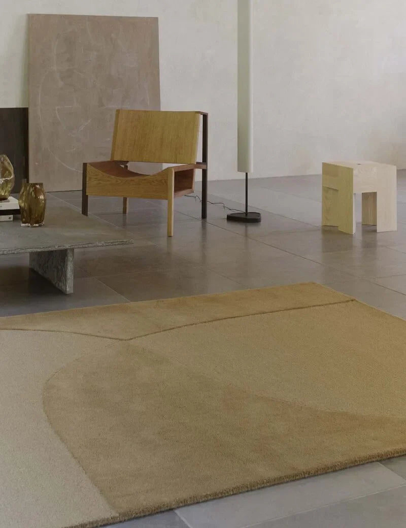 Großer rechteckiger Wollteppich in warmen Sand- und Goldtönen mit organischer Musterung in einem hellen, minimalistischen Wohnraum mit Holzmöbeln.