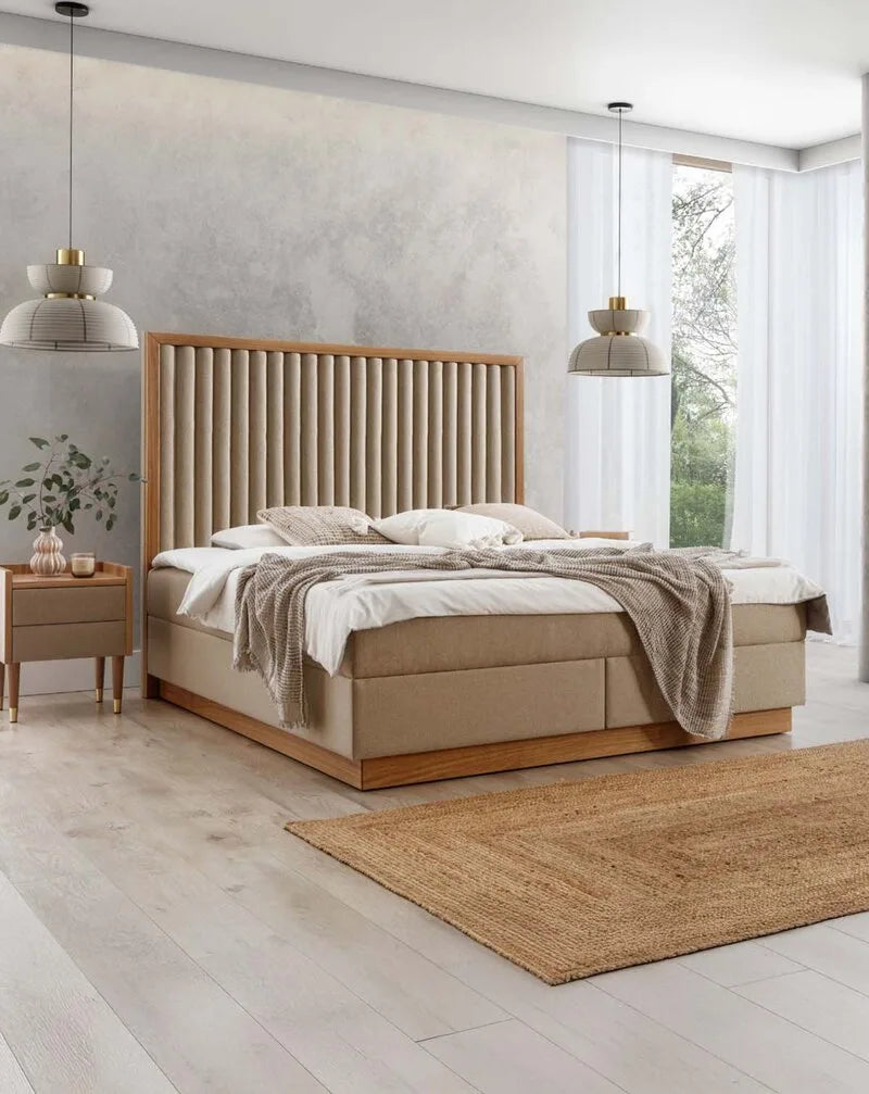 Modernes Boxspringbett mit gepolstertem, vertikal gestepptem Kopfteil in Beige und Holzrahmen, ergänzt durch Nachttisch und Jutéteppich im hellen Schlafzimmer.