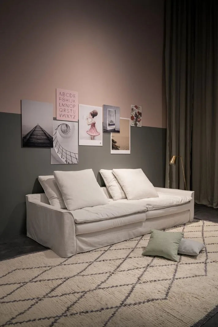 Weißes Hussensofa mit lockerem Bezug und großen Kissen vor zartrosa-graür Wand mit Bilderwand, Berberteppich und Stehlampe.