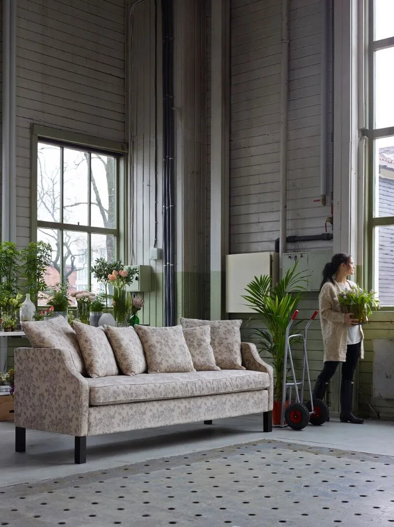 Gemustertes Landhaus-Sofa in beigen Blumenstoffen mit dunklen Holzfüßen vor grüner Holzwand in lichtdurchflutetem Loft mit Pflanzen.