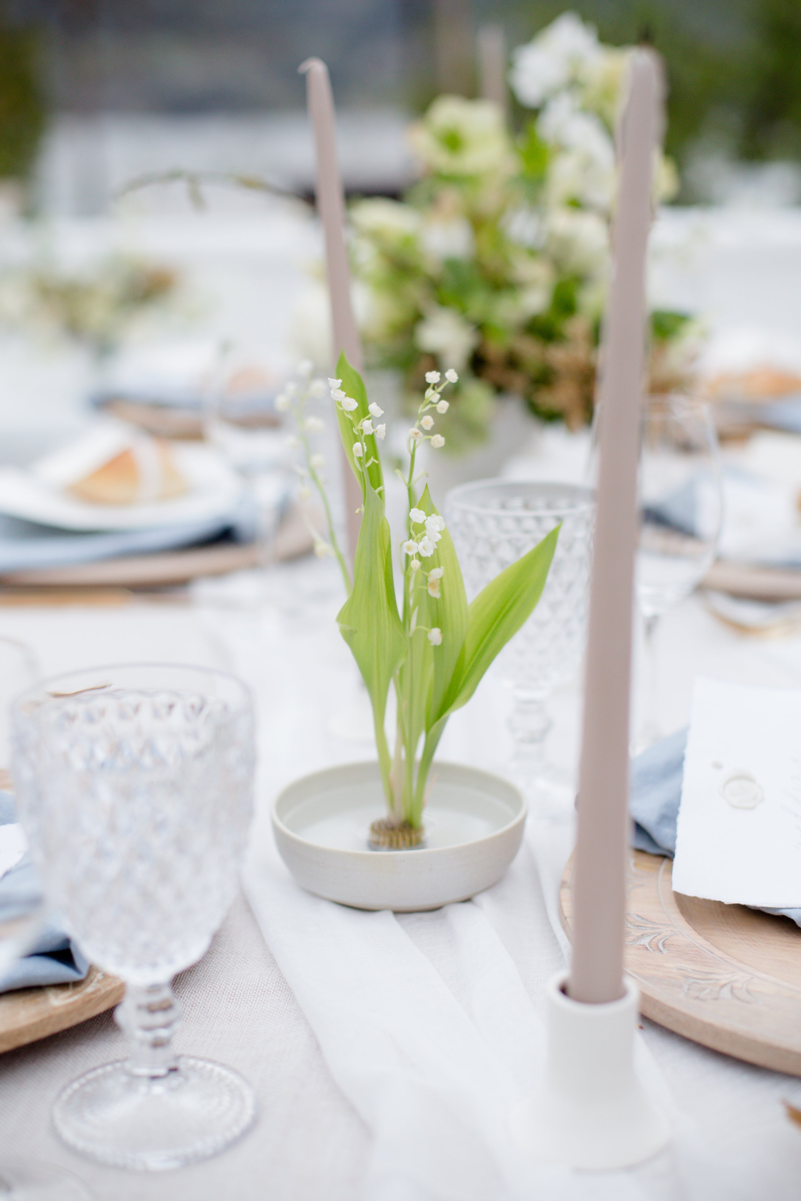 Eine Nahaufnahme eines elegant gedeckten Tisches mit Kristallgläsern, beigen Spitzkerzen und einer kleinen weißen Schale mit frischen Maiglöckchenblüten auf einer hellen Tischdecke mit dekorativen Tellern und Grünzeug im Hintergrund.